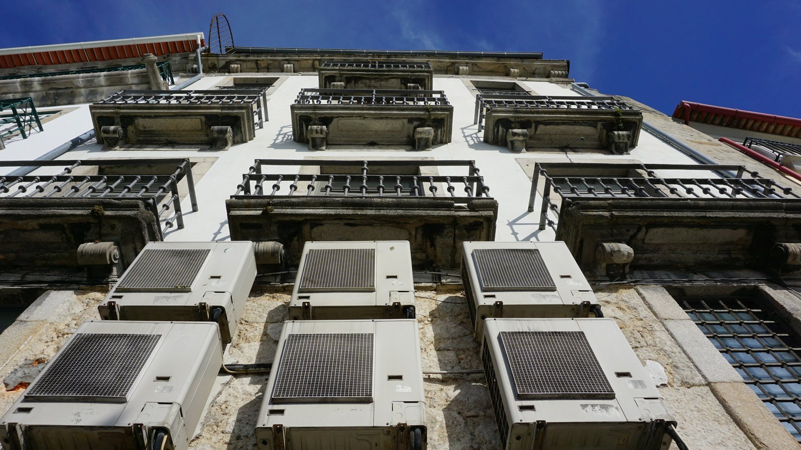 Seis unidades condensadoras de ar-condicionado fixadas na fachada de pedra de um prédio, vistas de baixo, sob varandas com gradil