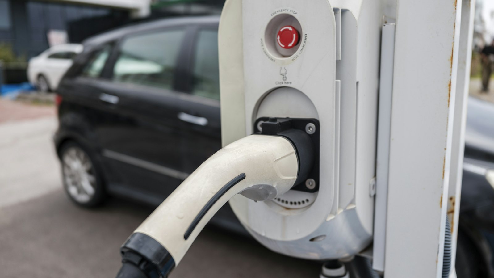 Estação de recarga de veículo elétrico em estacionamento externo, com o conector encaixado e o botão vermelho de parada de emergência no corpo do equipamento