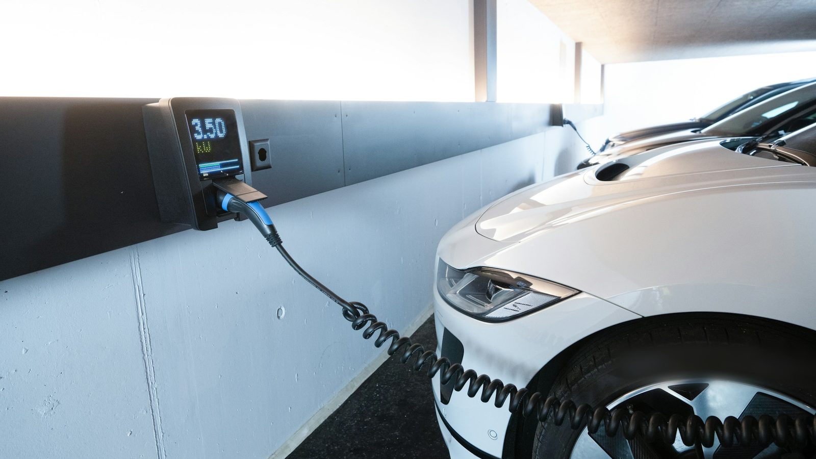 Carregador de veículo elétrico fixo na parede de uma garagem coberta, com o cabo conectado a um carro branco estacionado na vaga