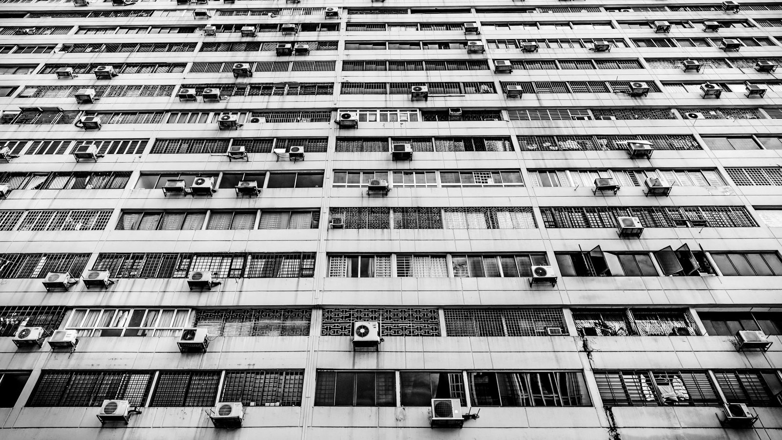 Fachada de um prédio residencial alto, vista de baixo, com dezenas de unidades condensadoras de ar-condicionado fixadas sob as janelas de cada andar