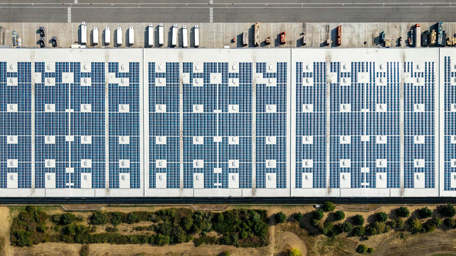 Vista aérea da cobertura de um galpão logístico tomada por módulos fotovoltaicos organizados em blocos, com faixas livres entre eles, docas de carga acima e vegetação abaixo