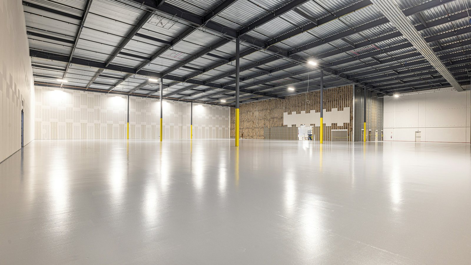 Interior de um galpão vazio, com piso de concreto liso, pilares metálicos de base amarela e cobertura de telhas metálicas com luminárias acesas