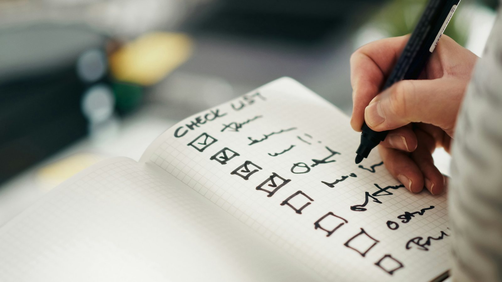 Mão com caneta preenchendo uma lista de verificação em um caderno, com parte dos itens marcada e parte ainda em aberto