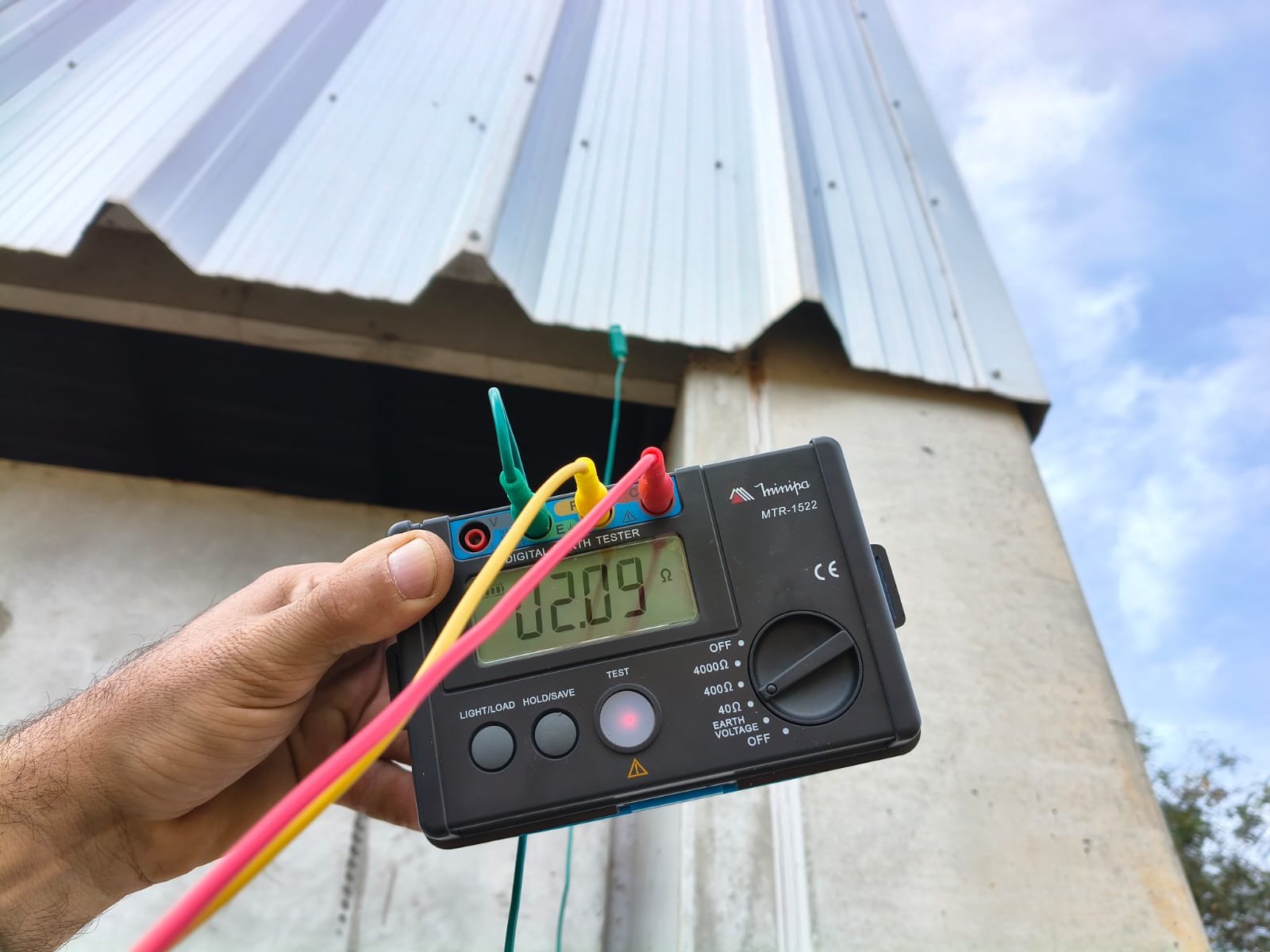 Terrômetro digital indicando 2,09 ohms na mão do técnico, com os cabos de ensaio ligados ao aterramento de um galpão de telha metálica