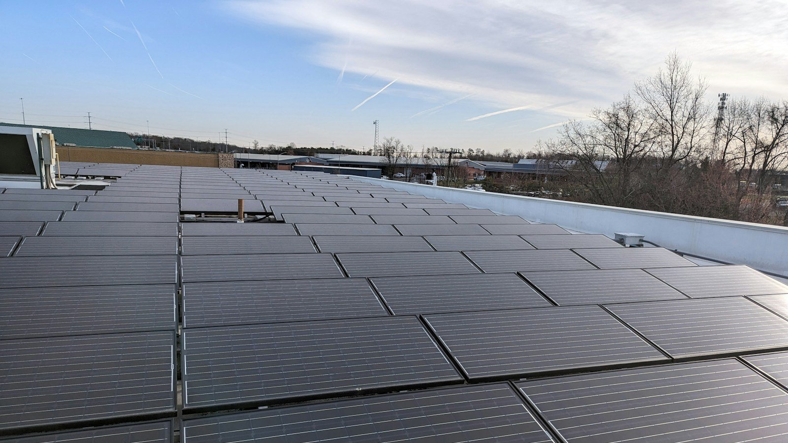 Fileiras de módulos fotovoltaicos sobre a laje de cobertura de uma edificação, junto à platibanda, sob céu azul com nuvens finas