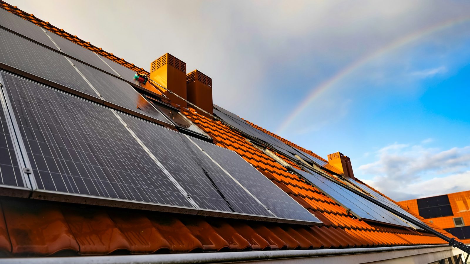 Fileiras de módulos fotovoltaicos sobre telhado de telhas cerâmicas molhadas, com céu ainda carregado e um arco-íris logo depois da chuva