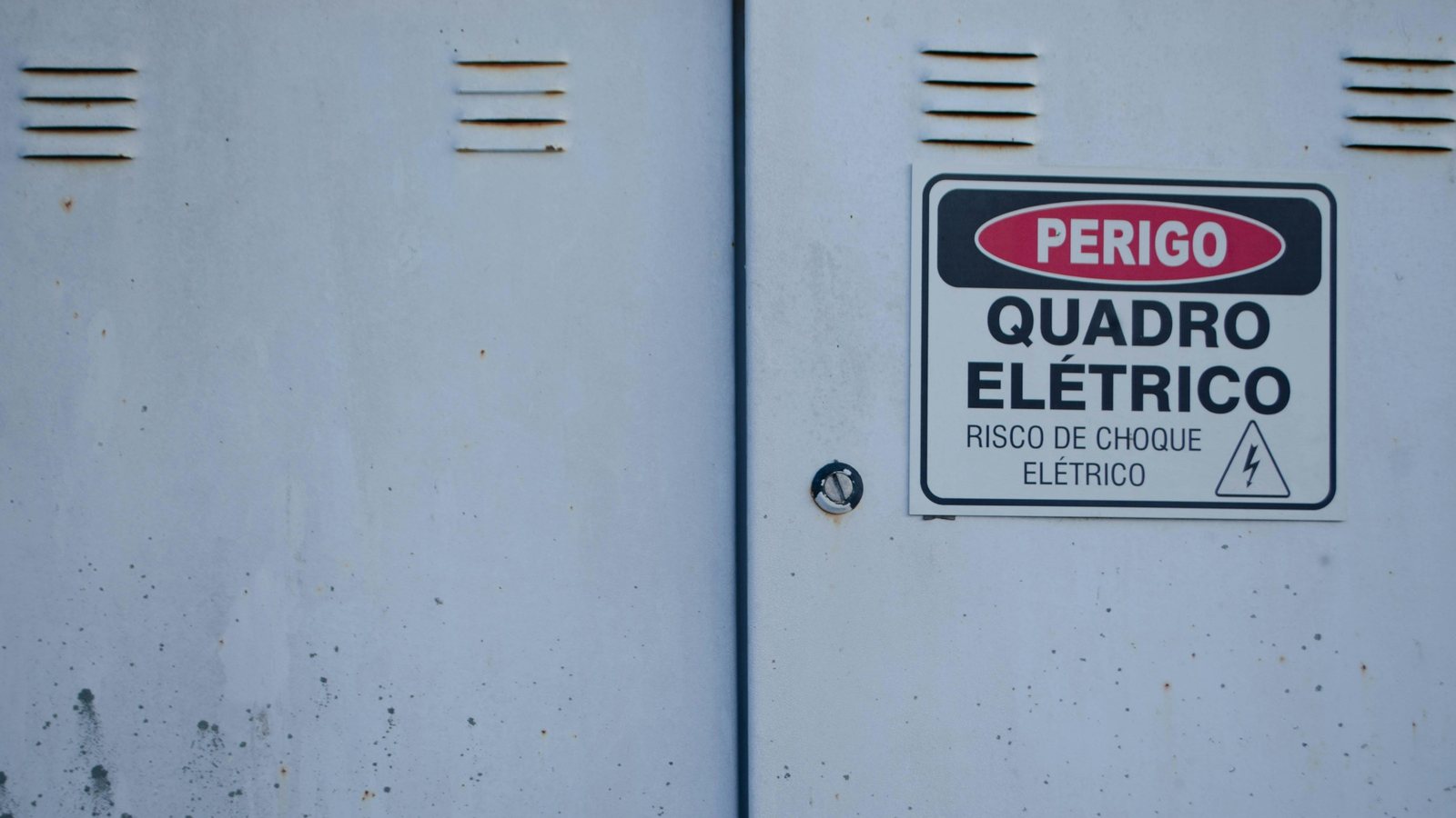 Portas metálicas fechadas de um quadro elétrico, com venezianas de ventilação e placa de advertência de perigo e risco de choque elétrico