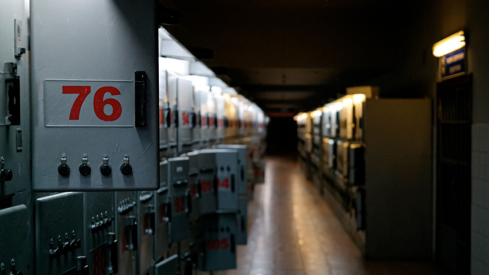 Corredor de sala elétrica com fileiras de painéis metálicos fechados e numerados, com botoeiras na porta e iluminação baixa