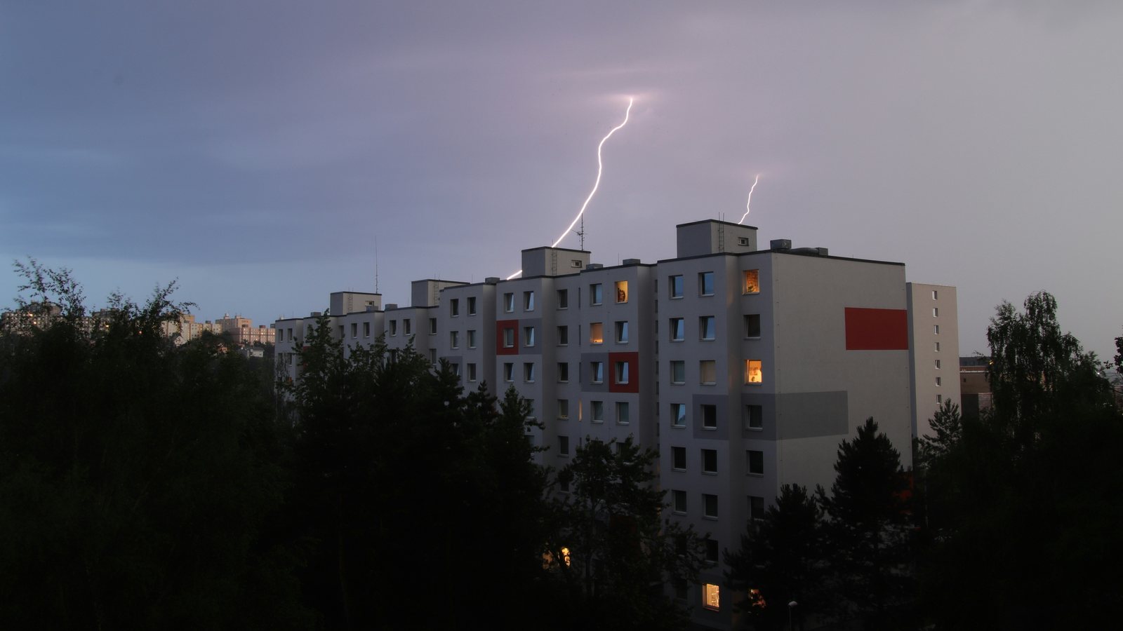 Descarga atmosférica descendo sobre a cobertura de um edifício residencial de vários andares ao entardecer, com casas de máquinas no telhado e árvores em primeiro plano
