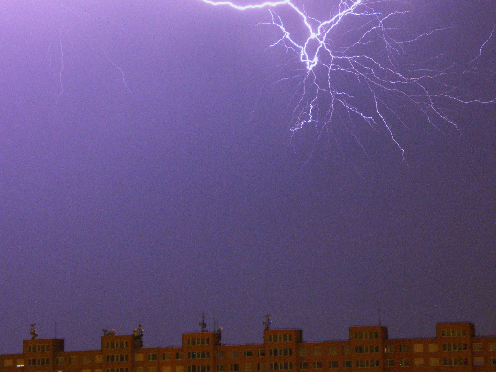 Descarga atmosférica ramificada no céu noturno arroxeado, acima da cobertura de um conjunto de prédios residenciais com antenas no topo