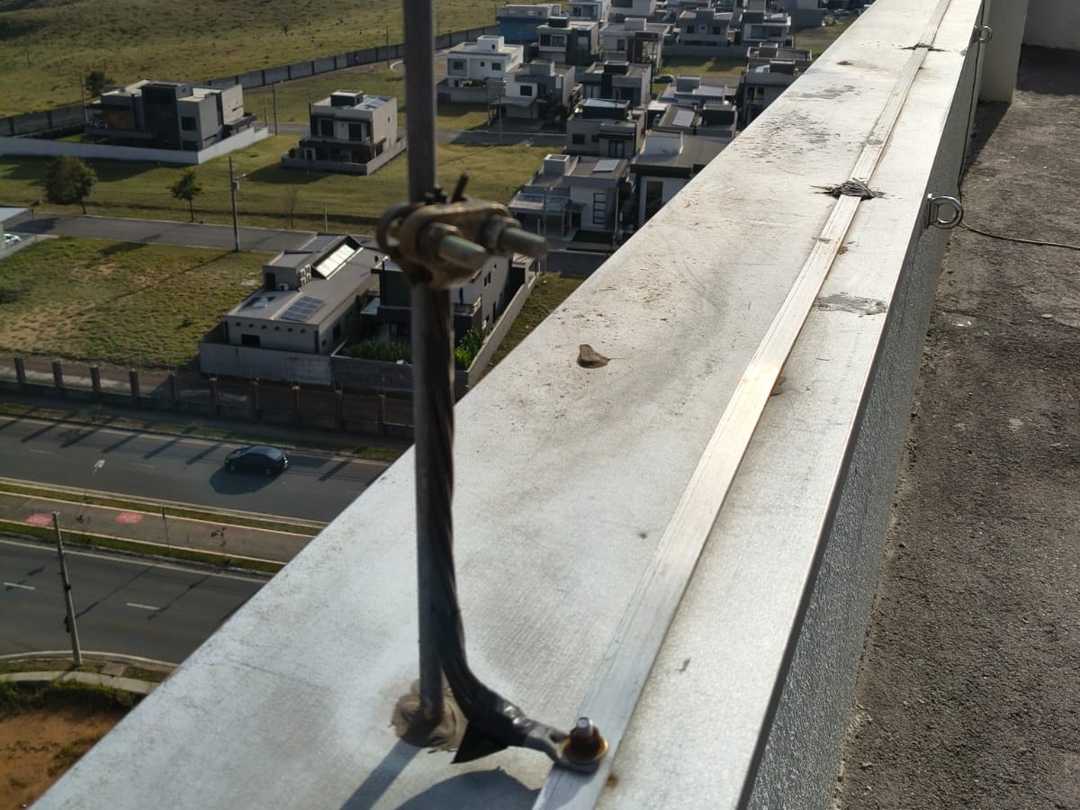 Haste captora do para-raios com oxidação, presa à platibanda da cobertura de um edifício, com um condomínio de casas e uma avenida ao fundo
