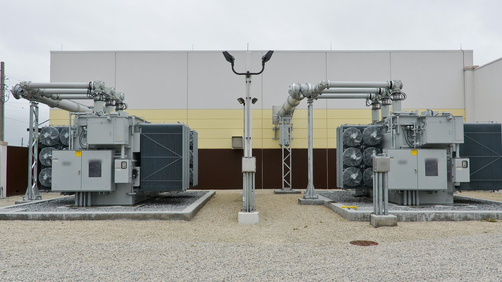 Dois transformadores de potência a óleo ao ar livre, cada um sobre base de concreto com leito de brita, ao lado da parede de uma edificação, com poste de iluminação entre eles