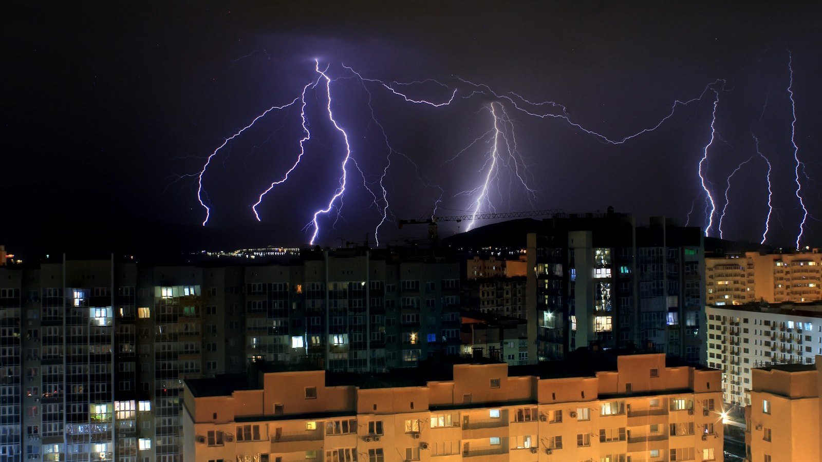 Vários raios no céu noturno atrás de prédios residenciais com janelas acesas, durante uma tempestade