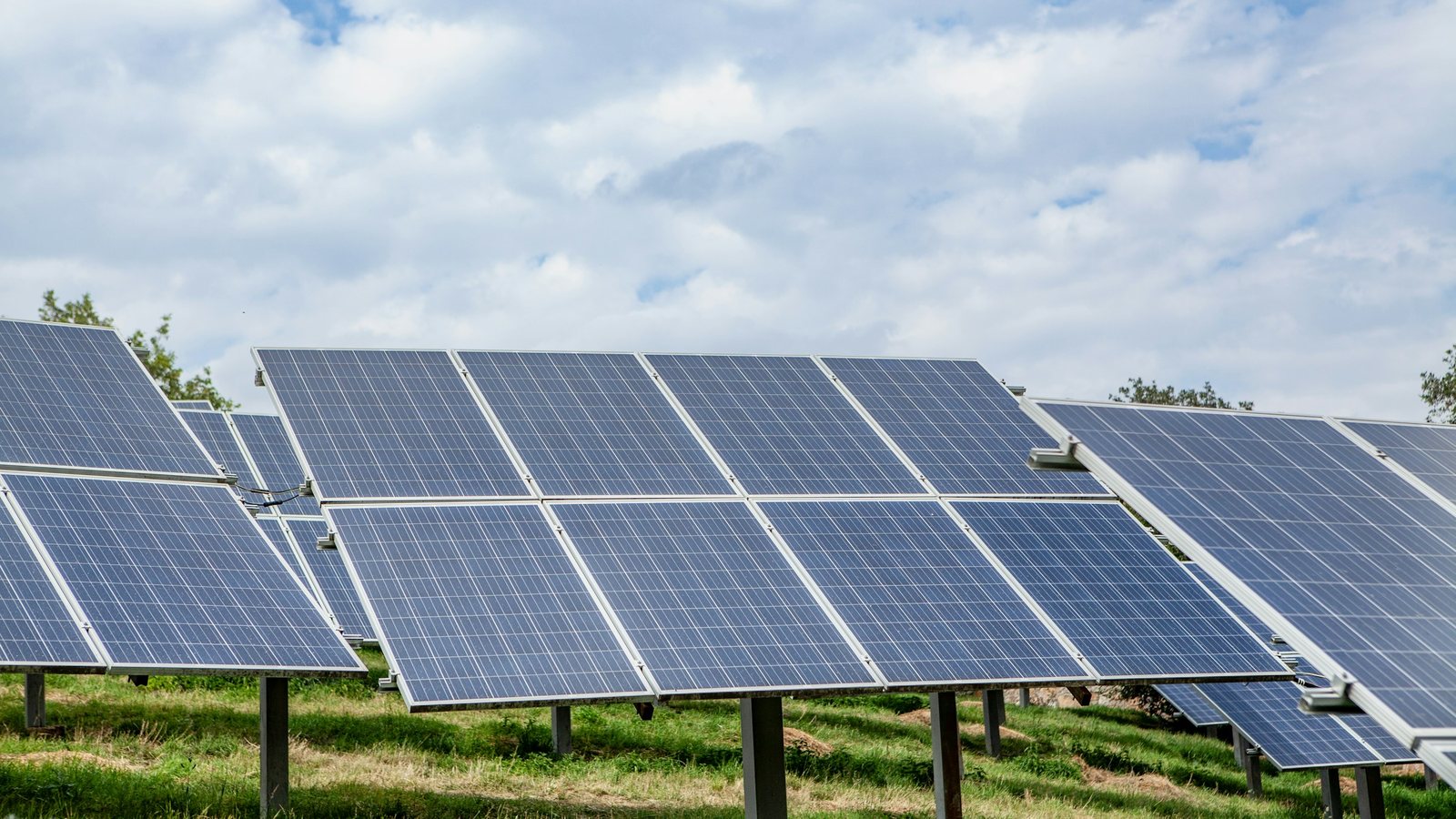 Fileiras de módulos fotovoltaicos montados sobre estruturas no solo, em terreno gramado de área rural, sob céu com nuvens